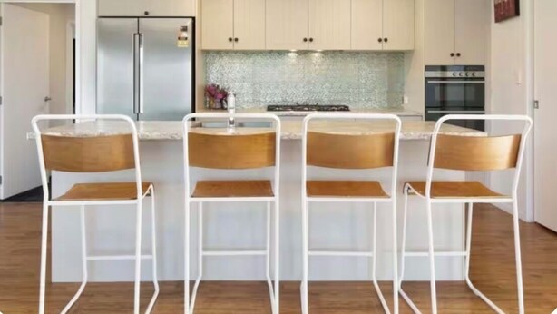 A modern kitchen with white cabinets, a stainless steel refrigerator, a built-in stove, and four bar stools.