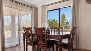A dining area with a wooden table and chairs, large windows with curtains, and a view of trees.