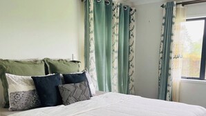 A neatly made bed with a white duvet, green patterned curtains, and a window with a view of trees.