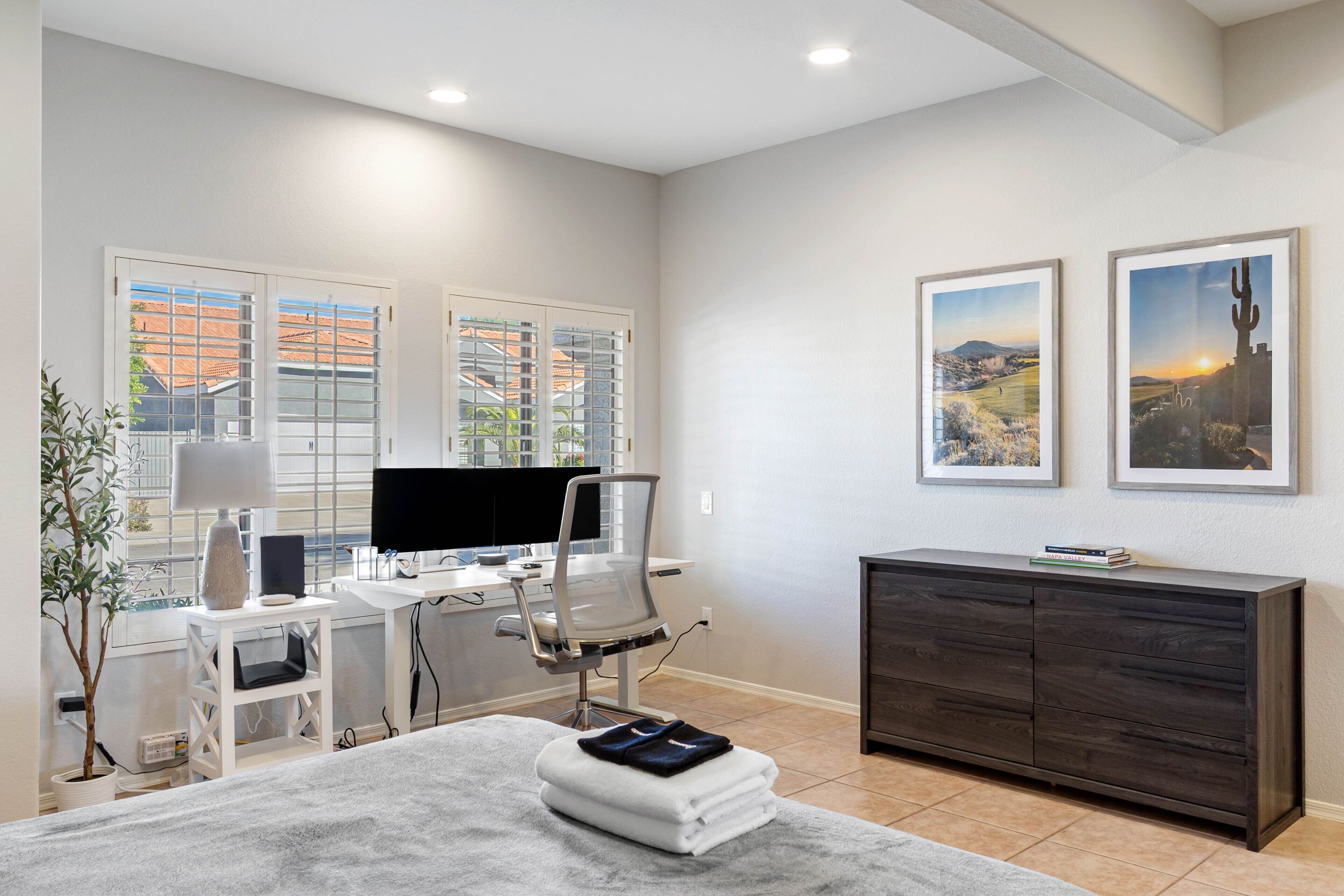 A modern bedroom with a desk, chair, and a large bed. There are two windows with blinds, a dark wooden dresser, and two framed pictures on the wall.