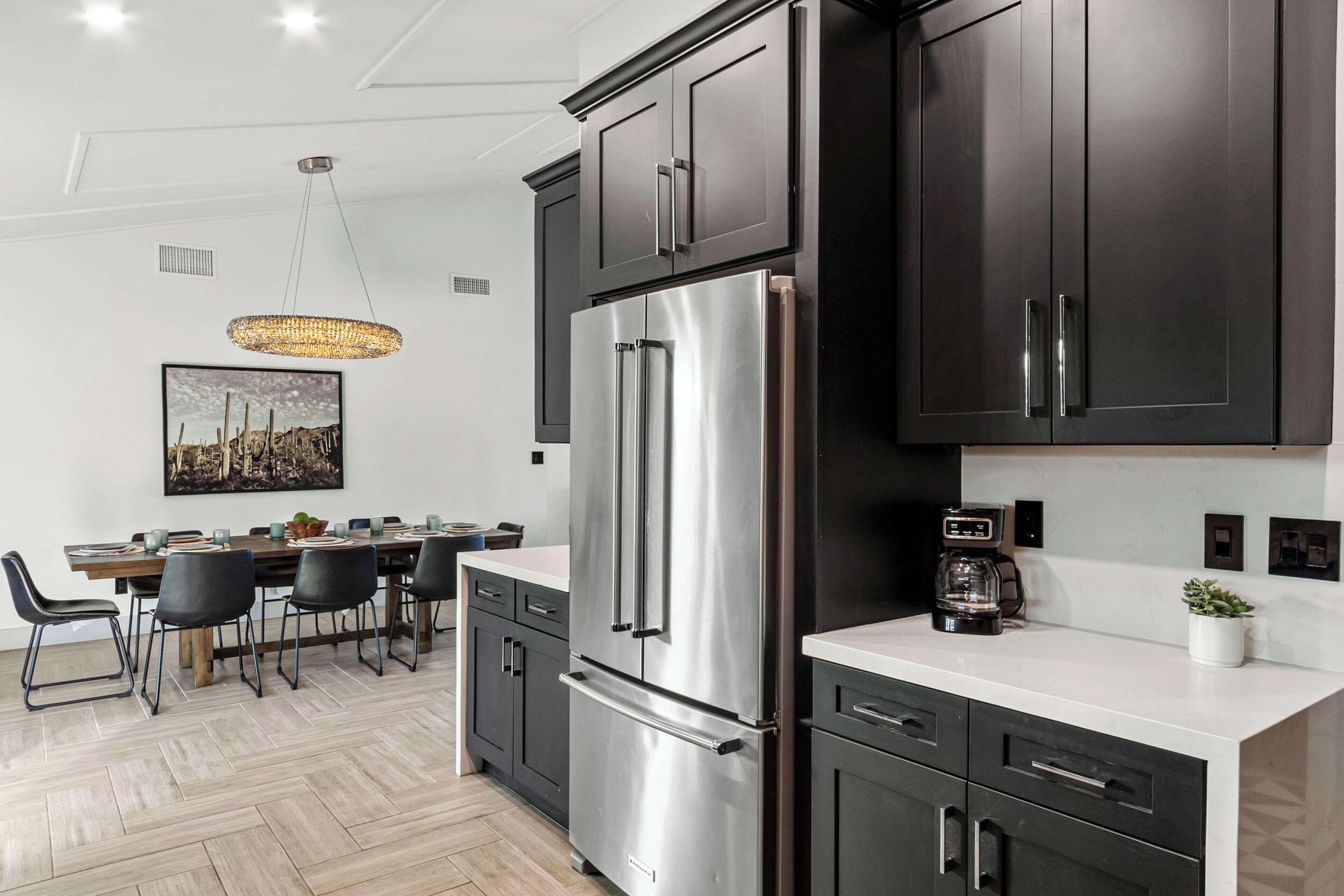 A modern kitchen with dark cabinets, stainless steel appliances, a dining area with chairs, and a hanging light fixture.