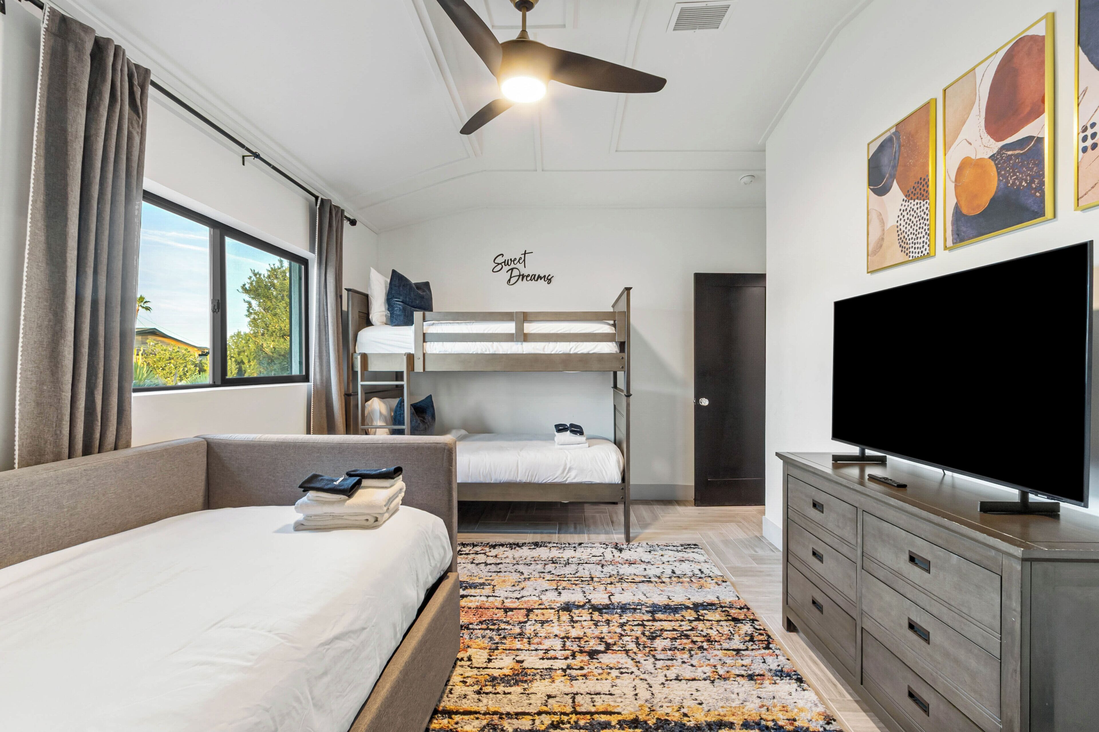 A modern hotel room with a bunk bed, a flat-screen TV, a ceiling fan, and a large window with curtains.