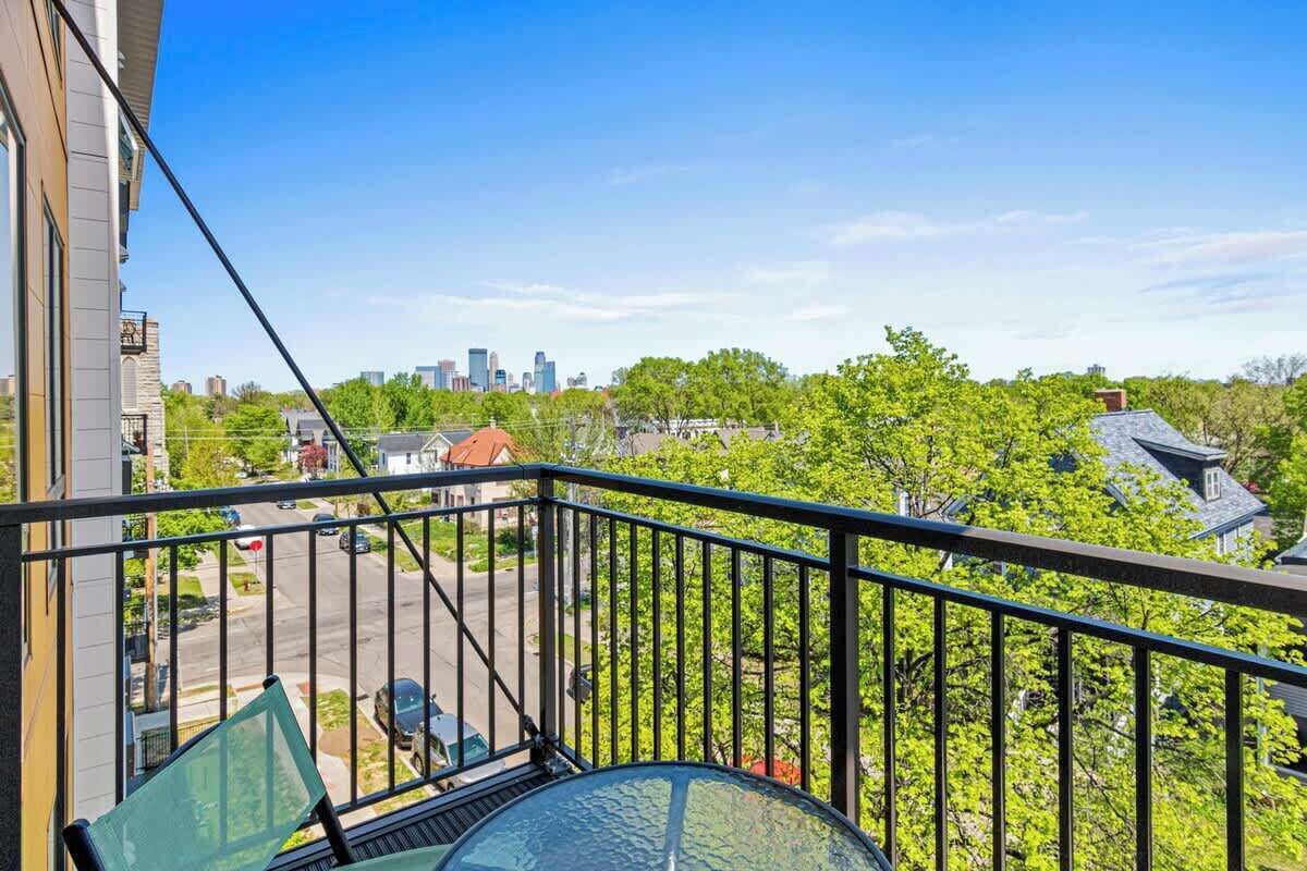 Un balcón con mesa y silla de vidrio, con vistas a una zona residencial con árboles y un horizonte urbano distante.