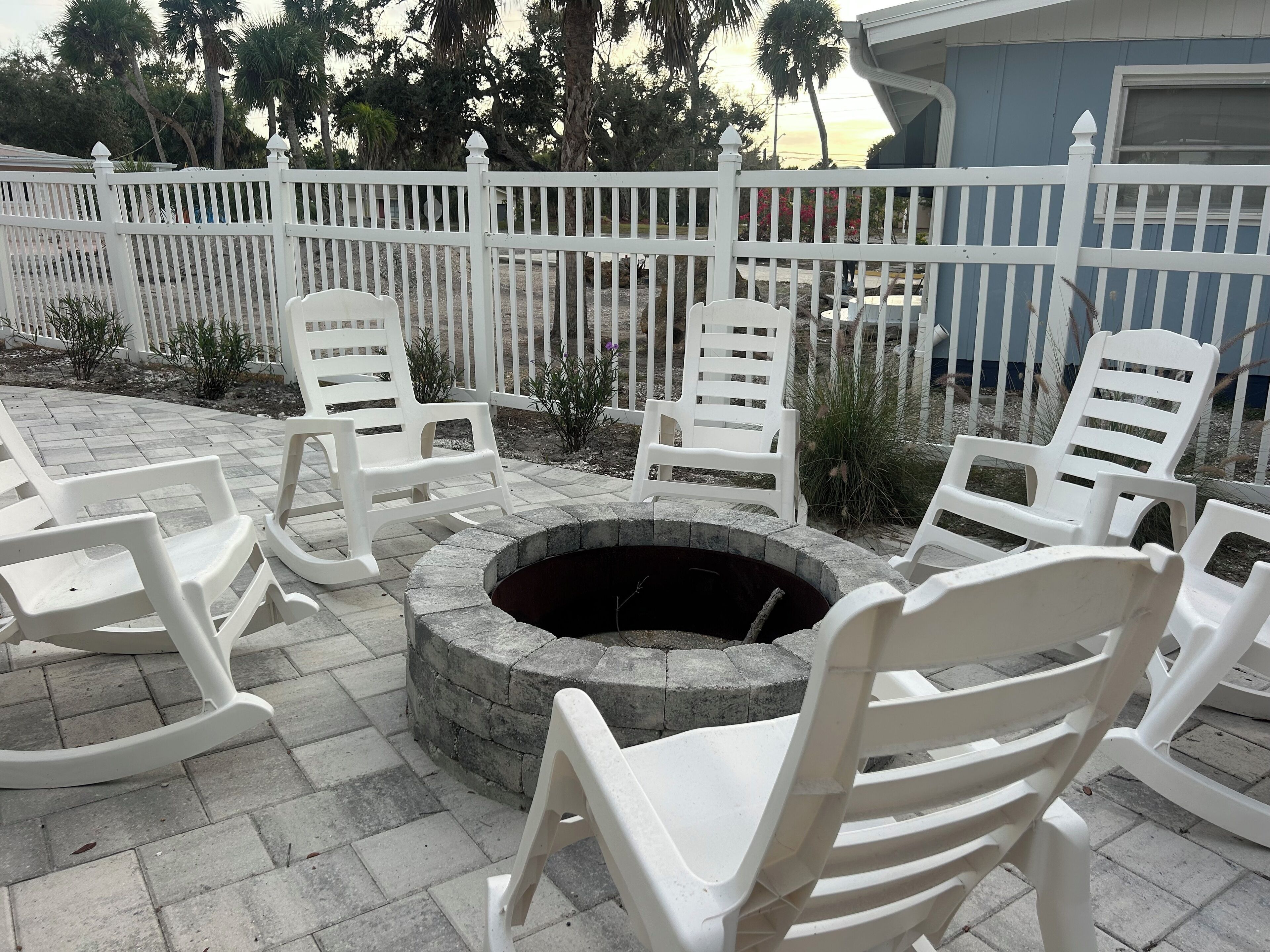 A fire pit surrounded by white plastic chairs, a white picket fence, and a building in the background.