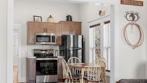 A compact kitchen with wooden cabinets, stainless steel appliances, and a dining area with wooden chairs. - The Bunkhouse | Downtown Branson Home Walkable to Landing & Convention Center (Branson)