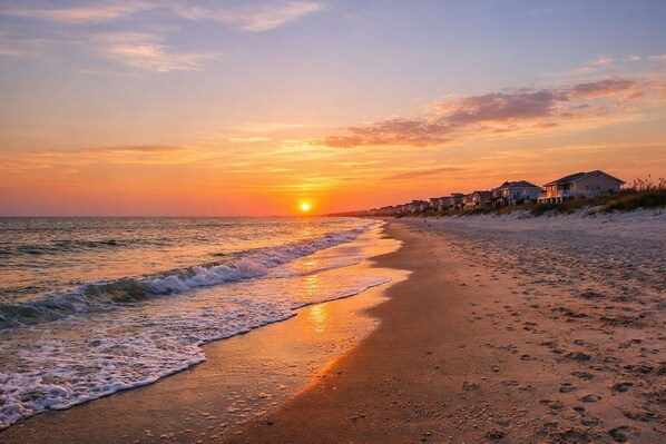 Ein Strand bei Sonnenuntergang, mit sandigem Ufer und Häusern am Wasser.