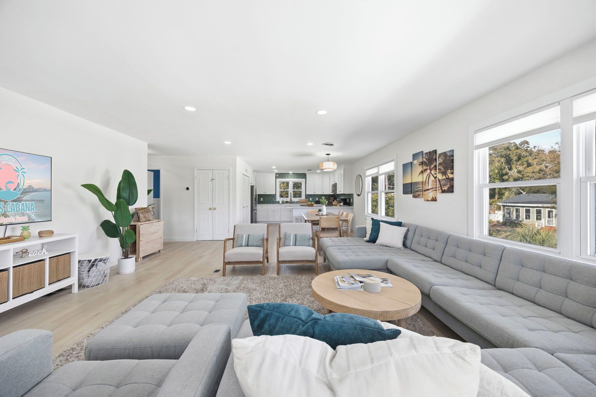 A modern living room with a large grey sectional sofa, a round coffee table, and a TV on a white stand. There is a kitchen in the background with white cabinets and a dining area with a table and chairs.