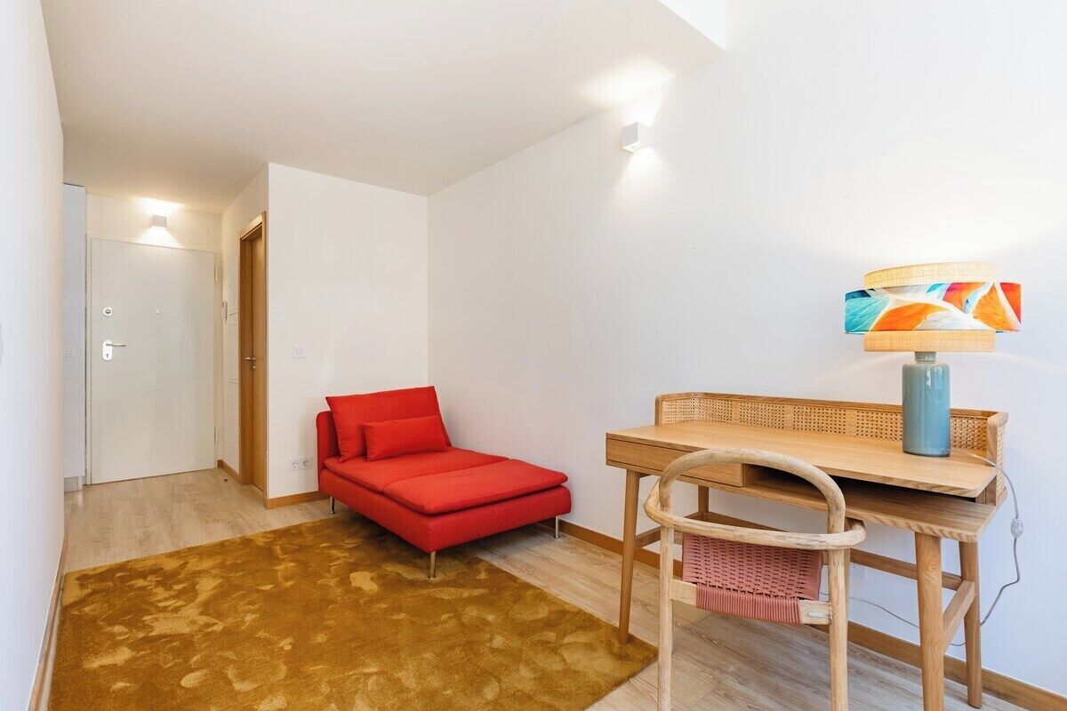 Une petite pièce avec un canapé rouge, une table en bois, une chaise, une lampe et un tapis marron.