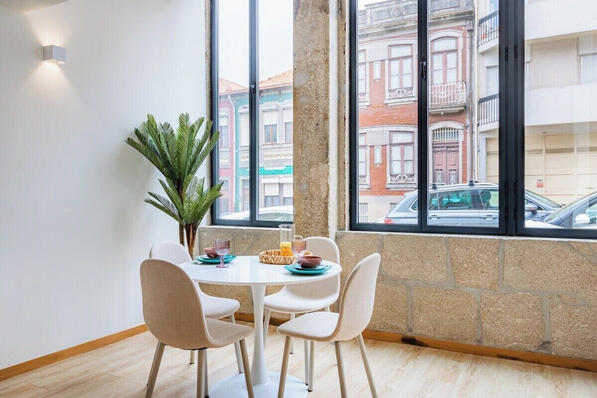 Un coin repas avec une table ronde, quatre chaises, une plante en pot et une vue sur les bâtiments à travers de grandes fenêtres.
