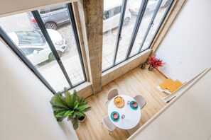 Ein Zimmer mit hohen Decken, einem großen Fenster mit Blick auf einen Parkplatz, einem runden Tisch mit Stühlen und einem Blumentopf.