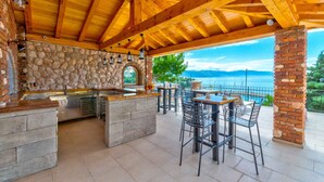 An outdoor kitchen area with stone walls, wooden beams, and a view of the sea. - Holiday apartment with wellness facilities and shared swimming pool (Novi Vinodolski)