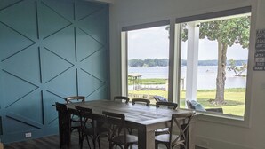 A dining area with a large table and chairs, a blue accent wall, and a view of a lake through large windows. - “Room for Everyone: Seven-Bedroom Lakefront Getaway on Lake Tillery” (Norwood)