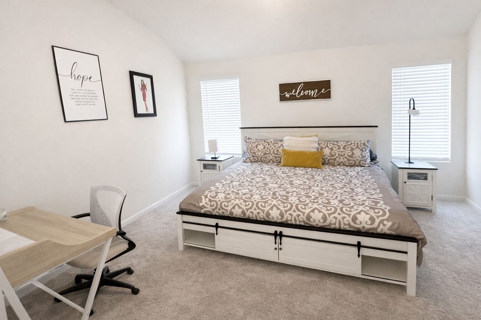 A bedroom with a bed, bedside tables, a desk, and a chair. There are two framed pictures on the wall and a 'welcome' sign above the bed.