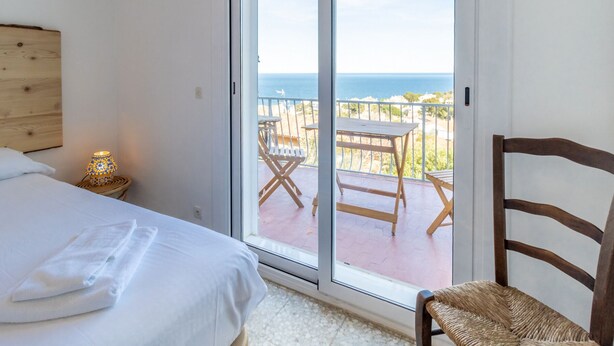 Ein Schlafzimmer mit einem Bett, einem Nachttisch und einer Lampe. Es gibt einen Balkon mit Tisch und Stühlen sowie Meerblick.