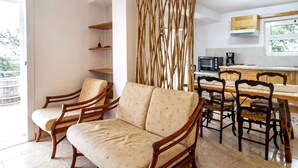 A living room with a sofa and chairs, a dining table with chairs, a kitchen area with appliances, and a view of greenery outside. - Fener de Dalt 86.2 - Apartment with views (Llança)