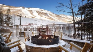 Un espacio al aire libre cubierto de nieve con un hogar de piedra, sillas de madera y vista a una montaña nevada.