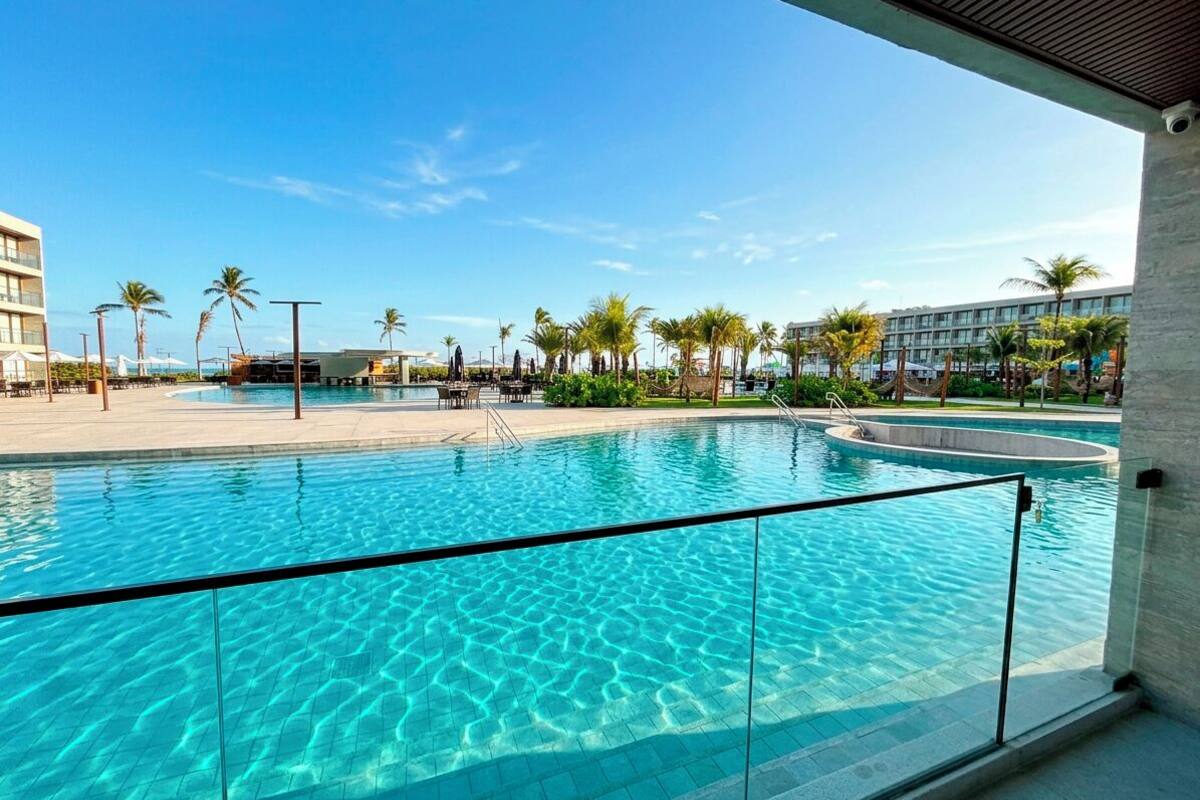 A clear swimming pool with a glass barrier, palm trees, and a modern building in the background.