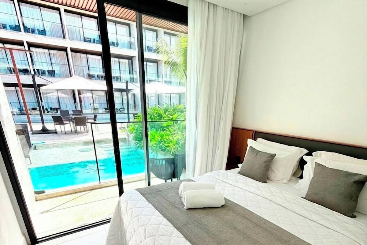 A hotel room with a bed, a view of a pool, and a balcony with chairs and umbrellas.
