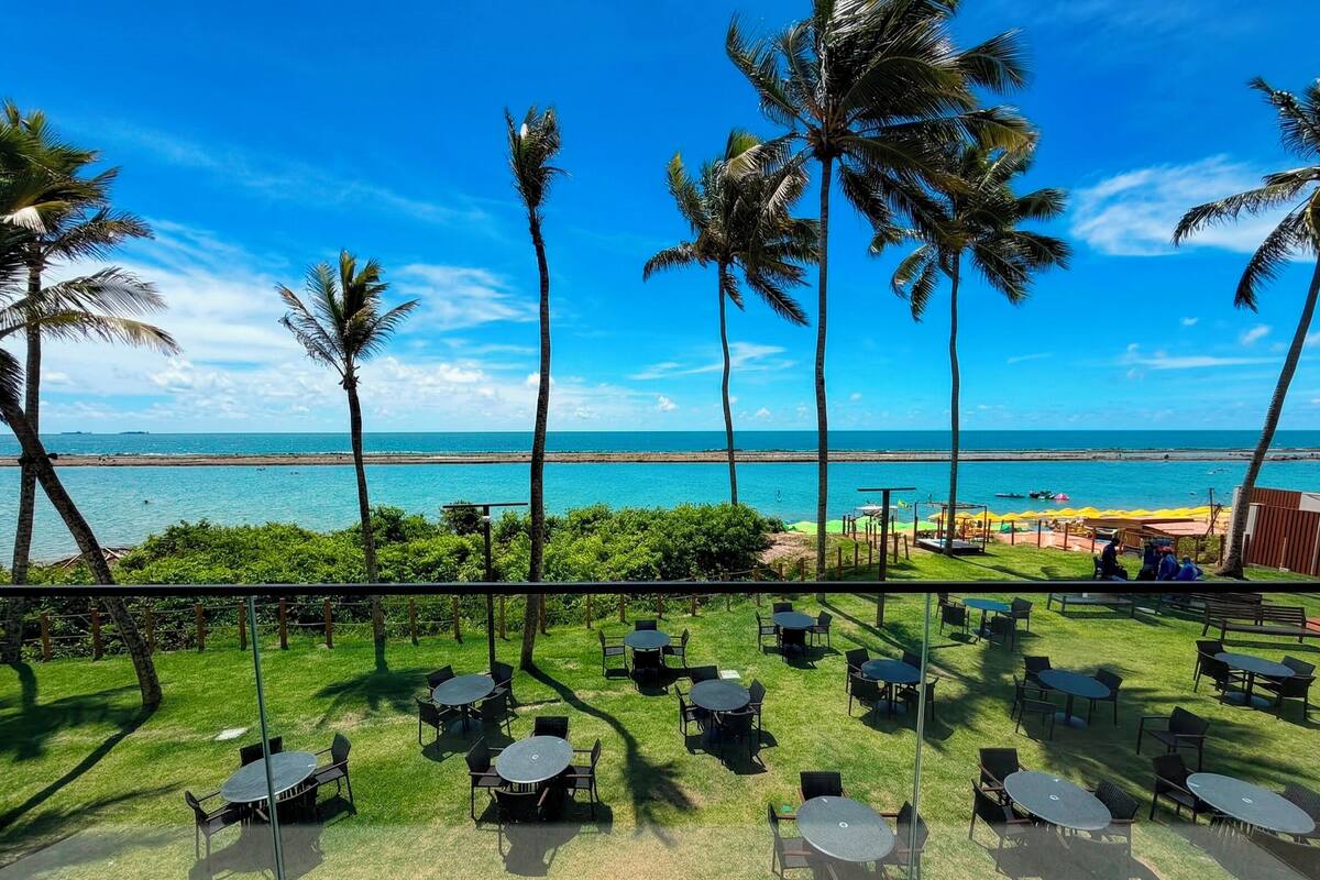 A terrace with tables and chairs overlooking a beach with palm trees and the ocean.