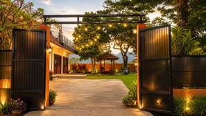 A gated entrance with a pathway leading to a building, surrounded by greenery and a tree with string lights. - Villa Feridys (Banjaran)