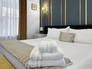 Ein Hotelzimmer mit einem ordentlich bezogenem Bett, einer Nachttischlampe, einer Wandleuchte und einem Fenster mit durchsichtigen Vorhängen.