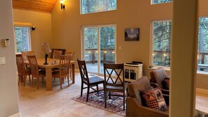 A spacious room with a dining area, a sofa, and large windows offering a view of the forest.