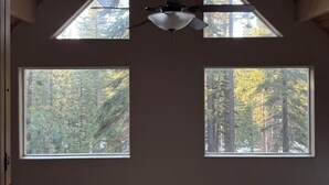 A room with a sloped ceiling, a ceiling fan, and three windows offering views of a forested area.