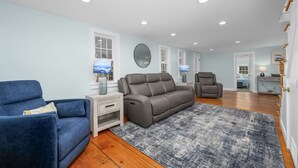 Un salón espacioso con un sofá gris, un sillón azul y otro sillón gris. Hay una mesa de centro de madera, una alfombra gris y un televisor en la pared.