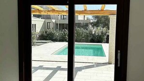 En vy genom en glasdörr in till ett poolområde med ett kaklat däck och en pergola ovanför.