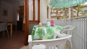 Et spiseområde med et bord dækket af en dug med et tropisk blad-mønster, hvide plastikstole og en grøn-stribet parasol.