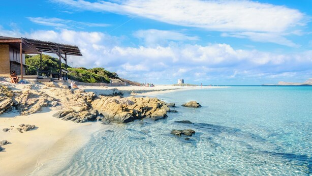 Ein Strand mit klarem Wasser, felsigem Ufer und einem Gebäude mit einer überdachten Fläche.