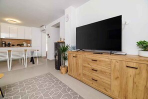 A modern living room with a large flat-screen TV on a wooden cabinet, a dining area with white chairs, and a kitchen in the background. - Holiday home with private pool and sea view (Selce)