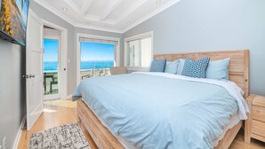 Un dormitorio con una cama grande, cabecera de madera, mesita de noche y vista a la playa a través de una puerta corredera.