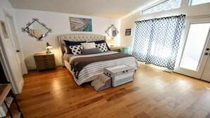 Un dormitorio con una cama grande, suelo de madera, una mesita de noche con lámpara, un aparador y una ventana con cortinas estampadas.