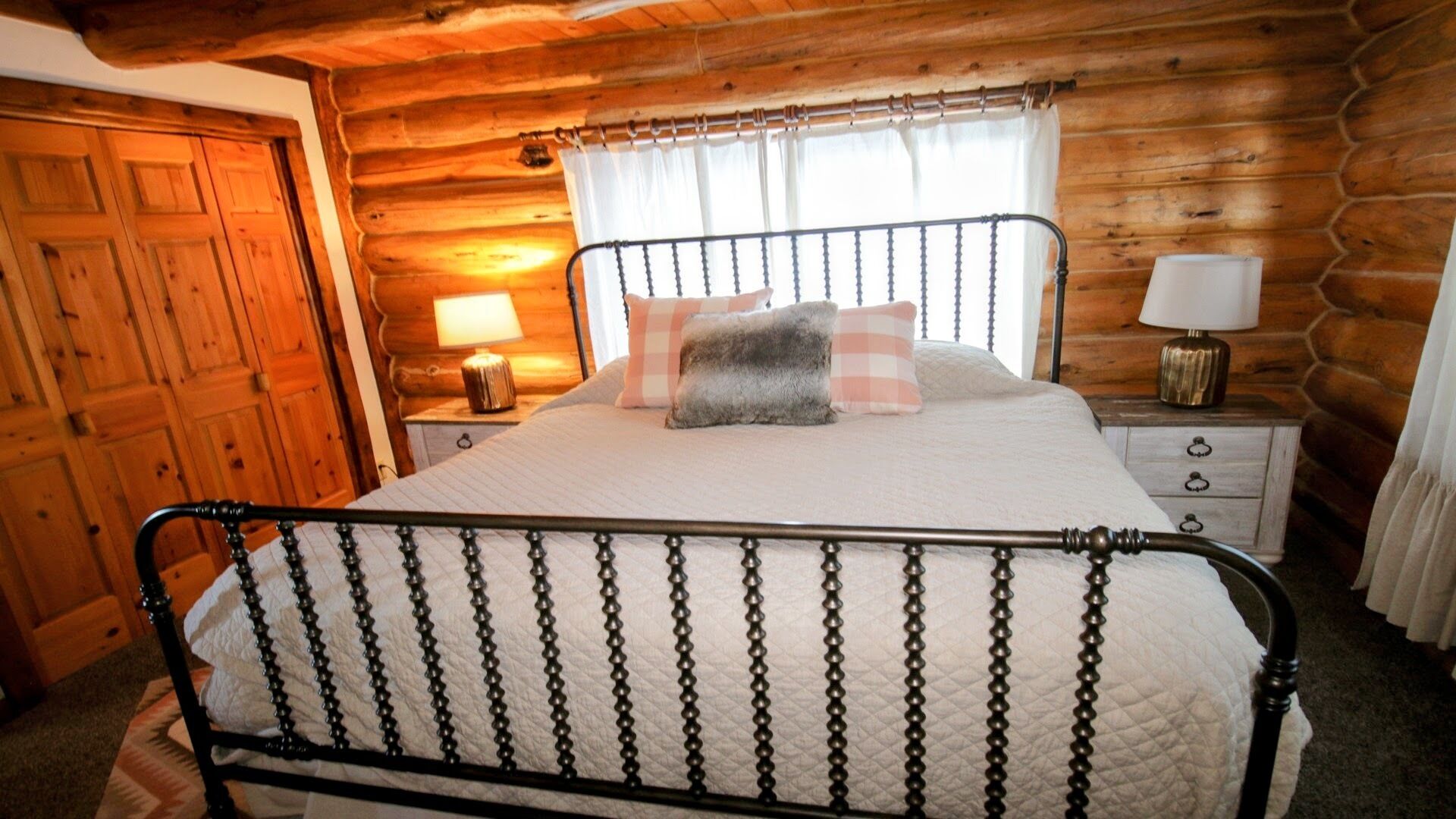 A wooden cabin bedroom with a metal bed frame, a nightstand with a lamp, and a window with curtains.
