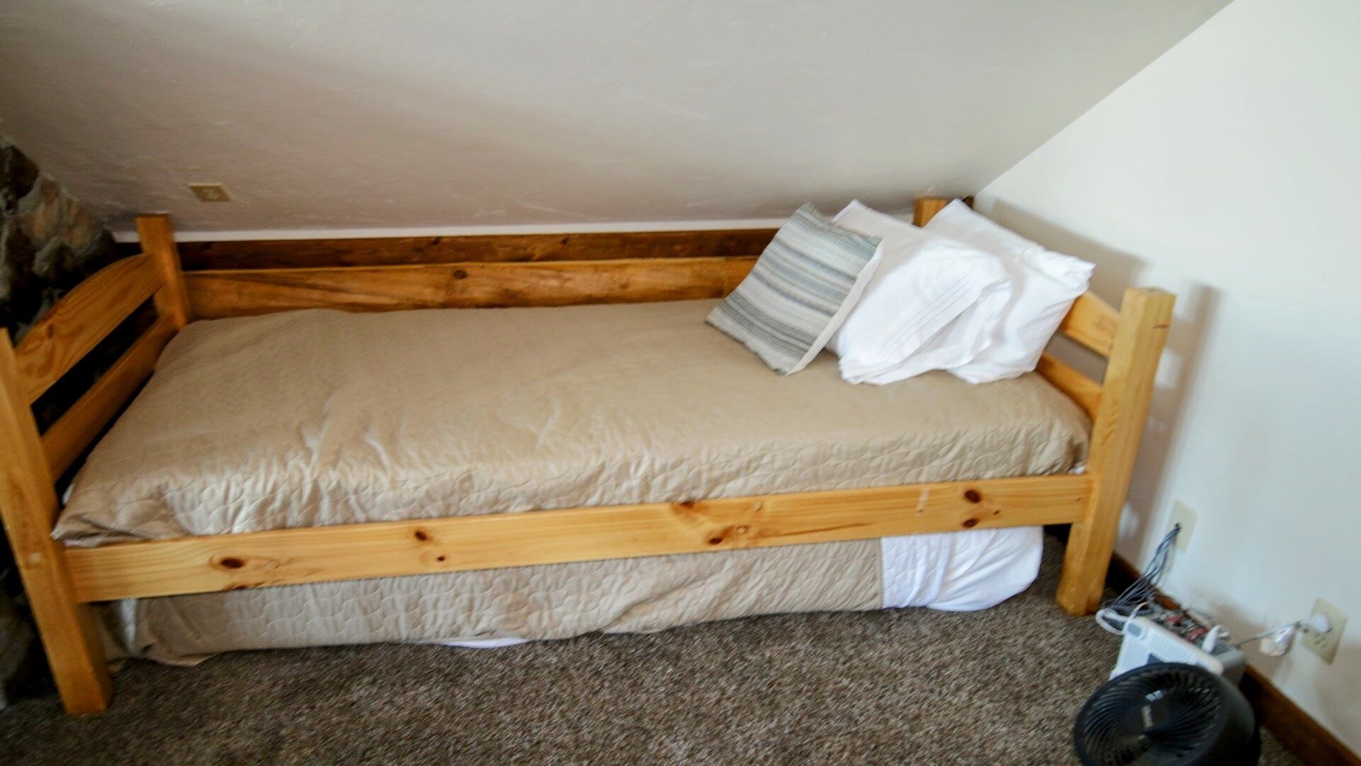 Un petit lit en bois, avec un matelas beige et des oreillers blancs, dans une pièce dont le sol est recouvert d’une moquette et dont un des murs est blanc.