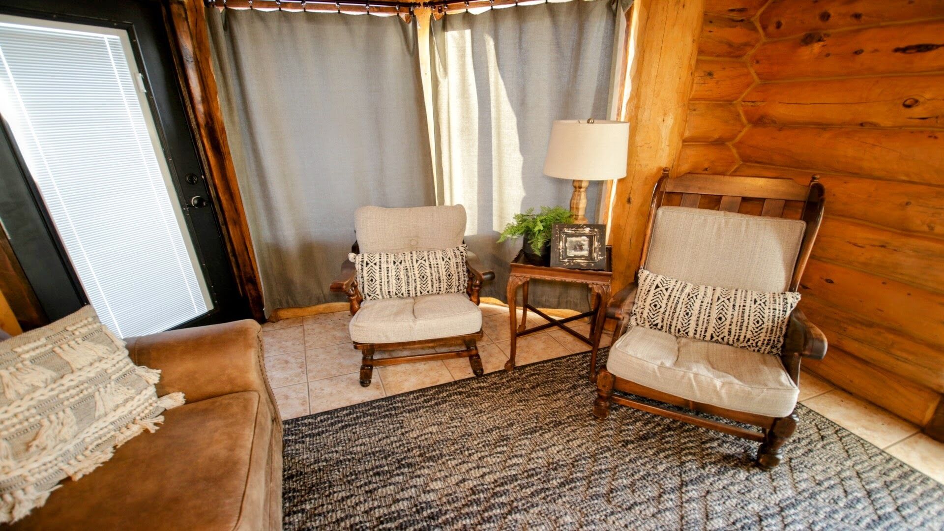 A cozy room with two wooden armchairs, a small table with a lamp, and a door with blinds.