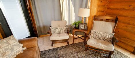 A cozy room with two wooden armchairs, a small table with a lamp, and a door with blinds.