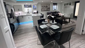 A modern kitchen with white cabinets, a dining area with a glass table and black chairs, and a living area with a black leather sofa and a mounted artwork. - Luxury 2BR/2BA Modern B Apartment in Ajax, Toronto (Ajax)