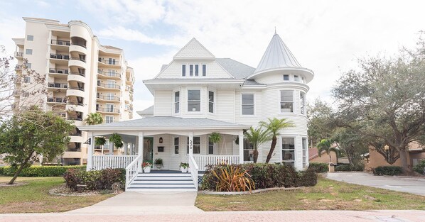 Une maison blanche à deux étages, dotée d’une véranda qui fait le tour de la maison, d’une tourelle et d’un jardin devant avec des plantes.