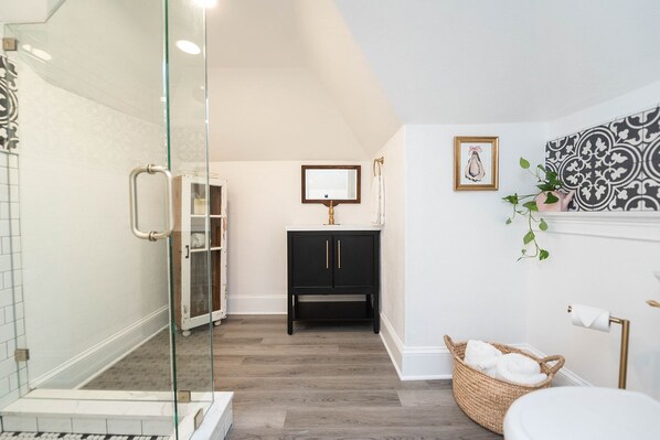 A modern bathroom with a glass shower enclosure, a black vanity with gold handles, a white sink, and a decorative wall with a framed picture.