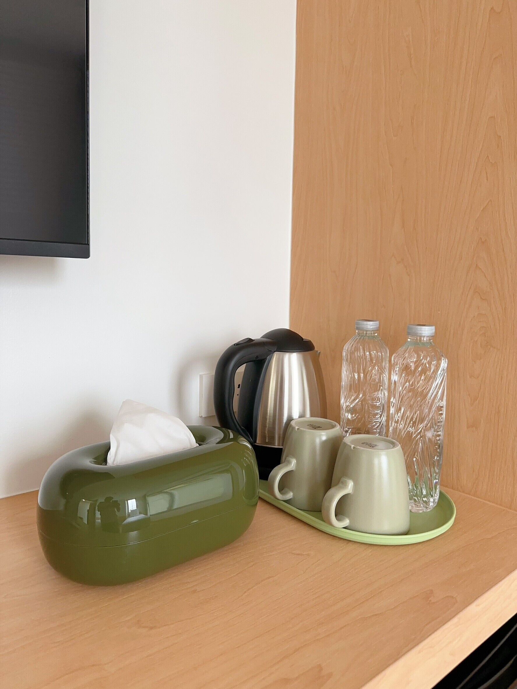 A neatly arranged kitchen area with a green tissue box, a kettle, two mugs, and two glass bottles on a wooden surface.