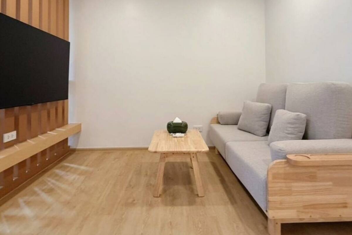 Uma sala de estar moderna com uma televisão de tela plana fixada em um painel de madeira, um sofá cinza, uma pequena mesa de centro de madeira e uma mesa de apoio de madeira.