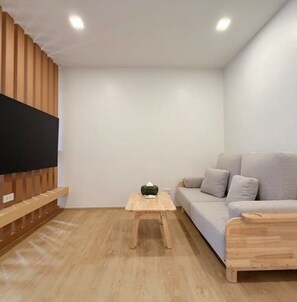 Un salon moderne avec un téléviseur à écran plat fixé sur un panneau en bois, un canapé gris, une petite table basse en bois et une table d’appoint en bois.