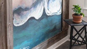 A large abstract painting on a wooden wall, a small potted plant on a black table, and a white door in the background.