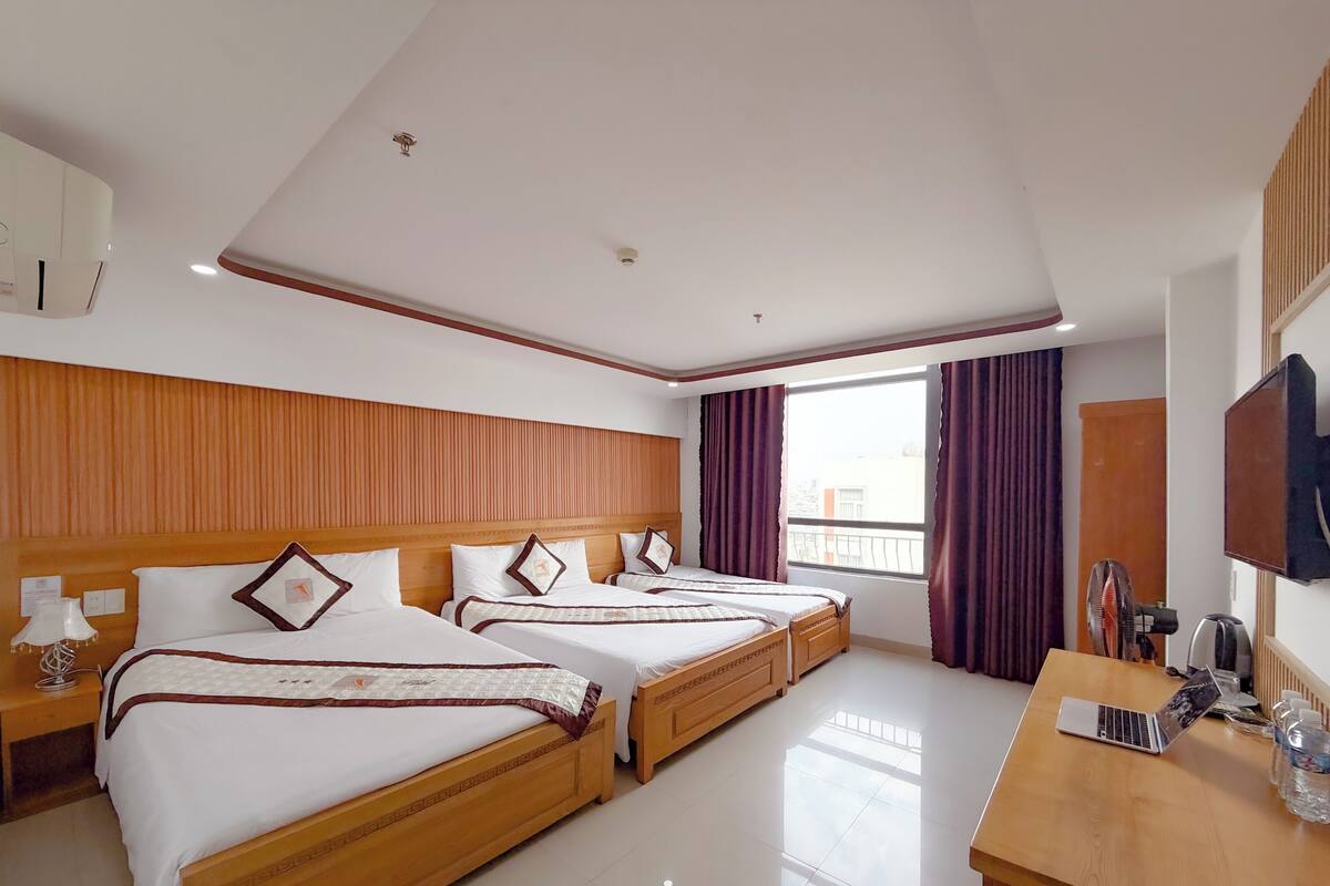 A hotel room with three wooden beds, a wooden desk, a television, and a window with curtains.