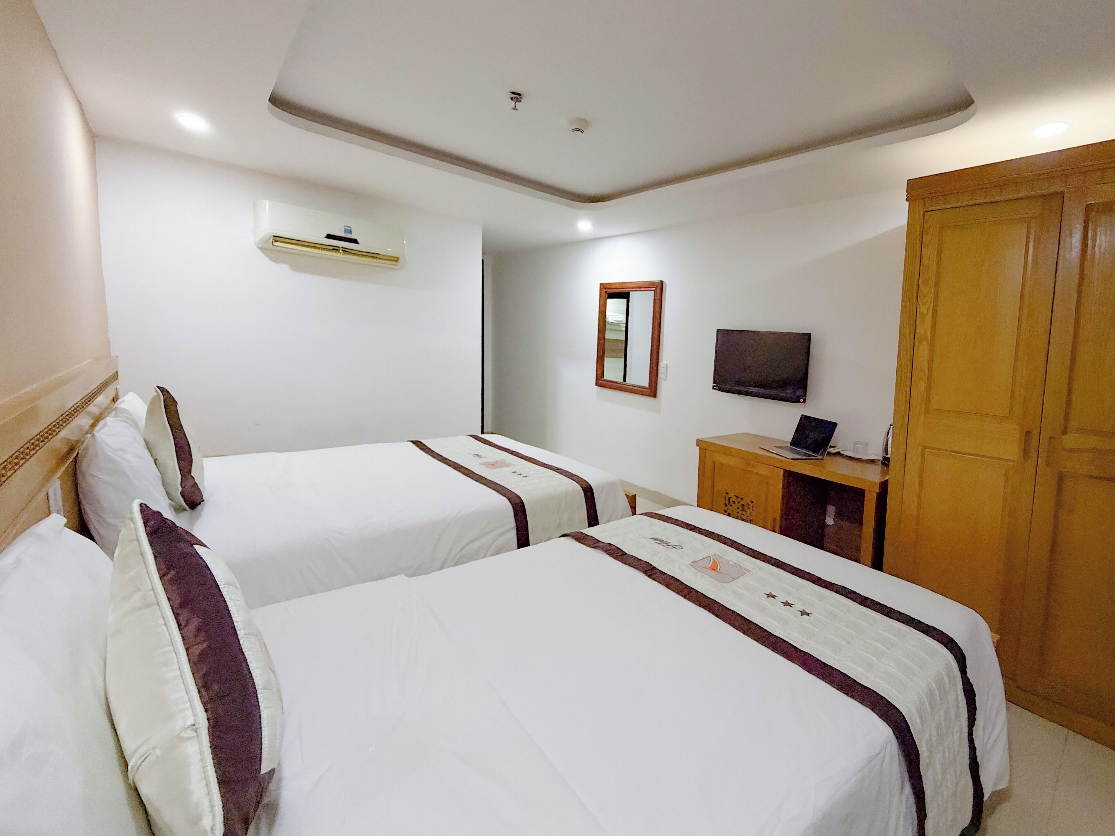 A hotel room with two beds, a TV, a wooden wardrobe, and a small desk with a laptop.