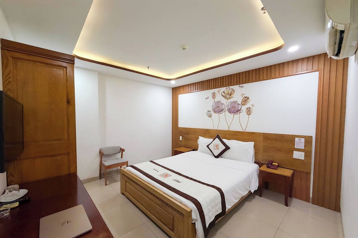 A bedroom with a wooden bed, a desk with a laptop, a chair, a nightstand, and a wall-mounted air conditioner.