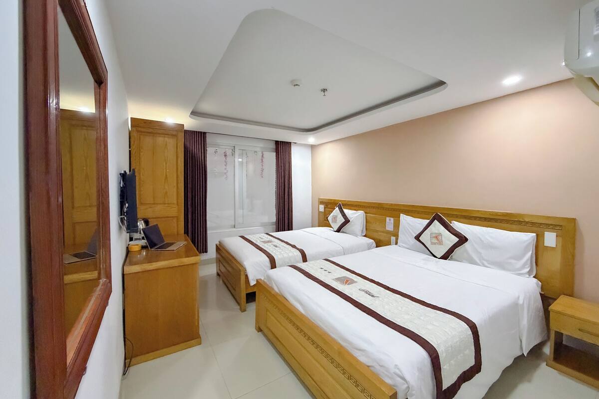 A hotel room with two beds, wooden headboards, a wooden nightstand, a desk with a chair, a mirror, and a ceiling light fixture.