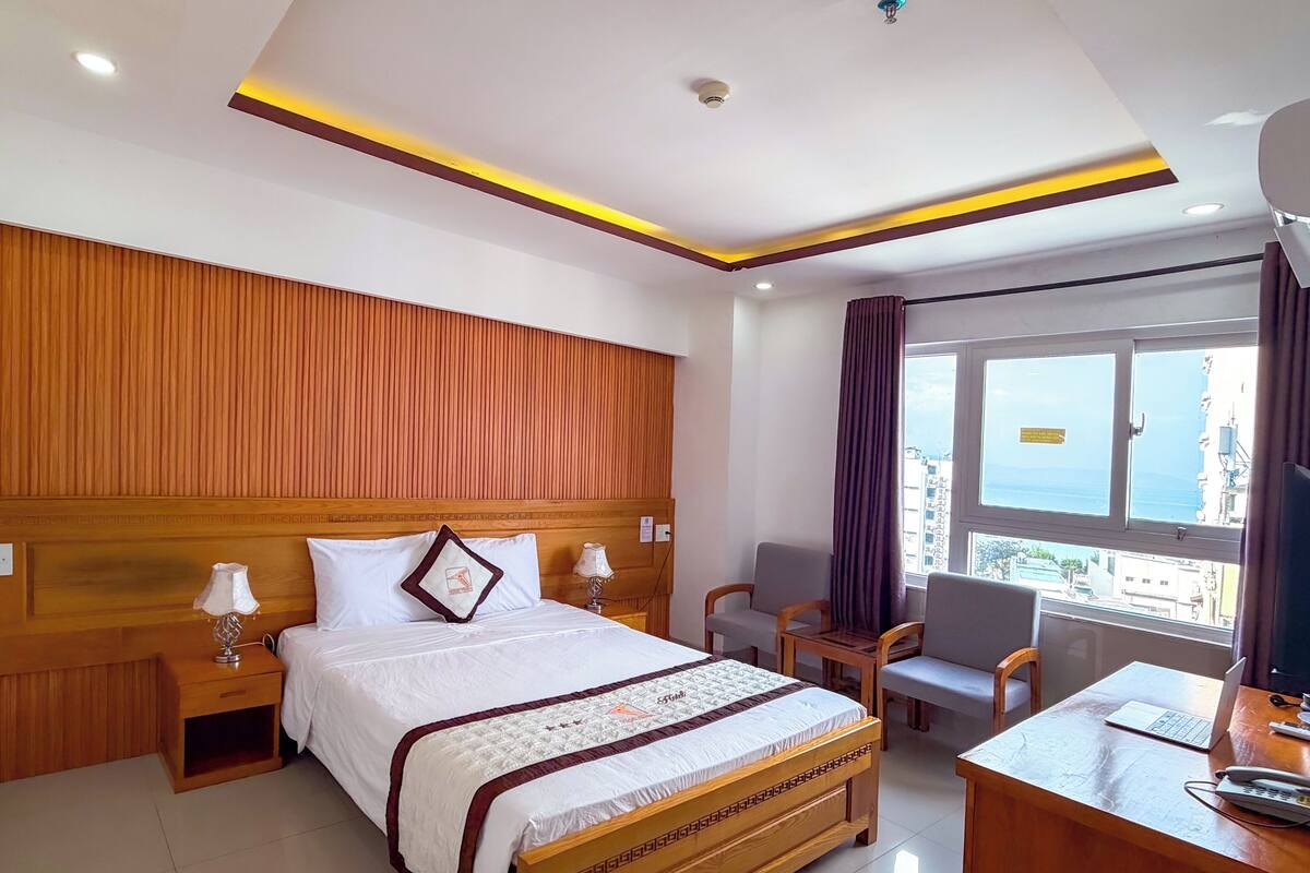 A hotel room with a wooden headboard, a bed with a white and brown bedspread, a desk with a laptop, a chair, and a view of the city through a window.
